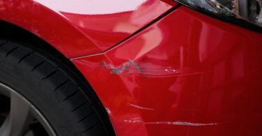 A close-up showing the scratched and chipped paint on a red car, focusing on the area around one of the rear tires.