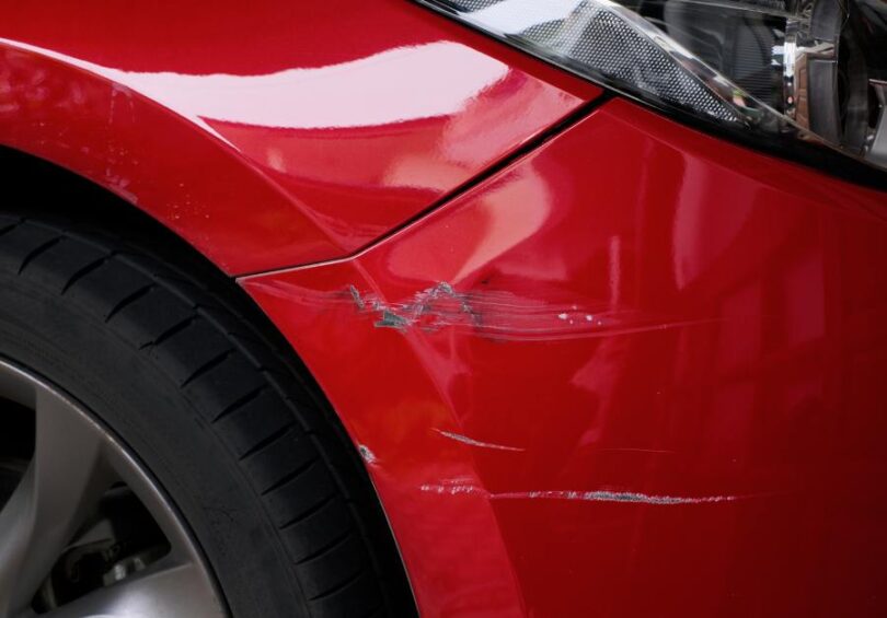 A close-up showing the scratched and chipped paint on a red car, focusing on the area around one of the rear tires.