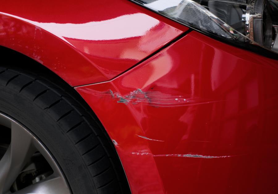 A close-up showing the scratched and chipped paint on a red car, focusing on the area around one of the rear tires.
