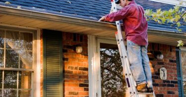 A man wearing a red button-down shirt, jeans, and a baseball cap is standing on a ladder cleaning gutters.
