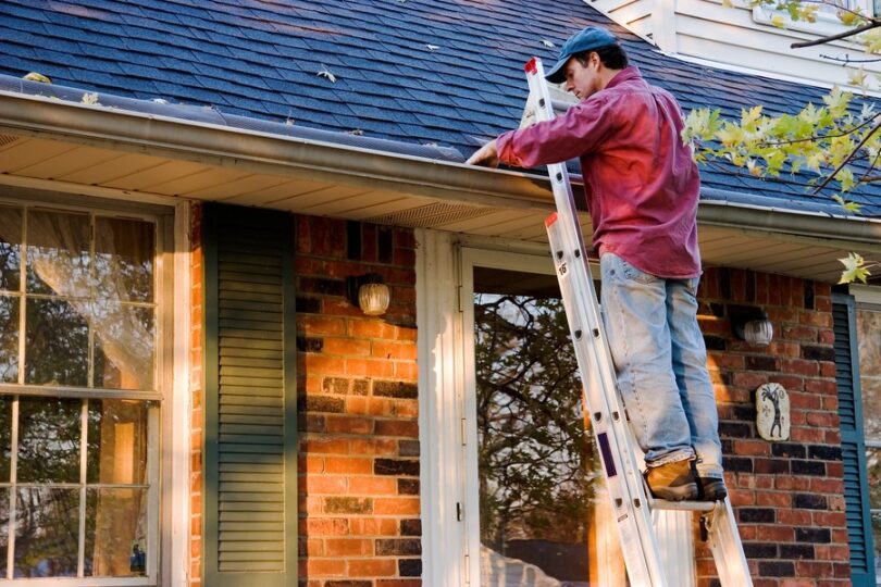 A man wearing a red button-down shirt, jeans, and a baseball cap is standing on a ladder cleaning gutters.