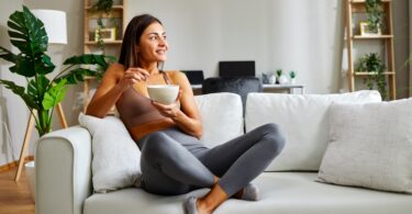 A young athletic woman is eating a bowl of healthy breakfast at home, sitting on the sofa and looking out the window.
