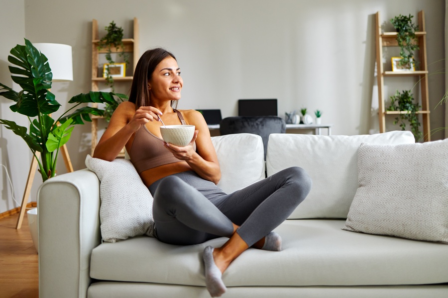 A young athletic woman is eating a bowl of healthy breakfast at home, sitting on the sofa and looking out the window.