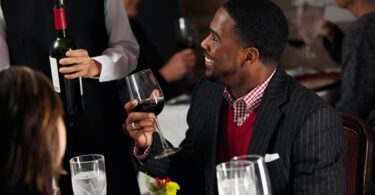 A well-dressed man in a sport coat sits at a table in a restaurant and holds a glass of wine. He smiles at the waiter.