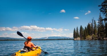 A person wearing orange clothing paddling a yellow kayak across a lake surrounded by a forest on a sunny day.