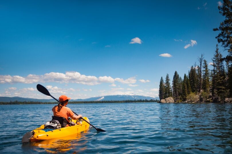 A person wearing orange clothing paddling a yellow kayak across a lake surrounded by a forest on a sunny day.