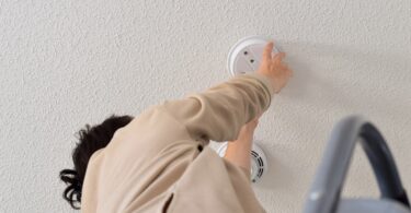 A person on a ladder installs a smoke detector on a textured white ceiling, emphasizing home safety and maintenance.
