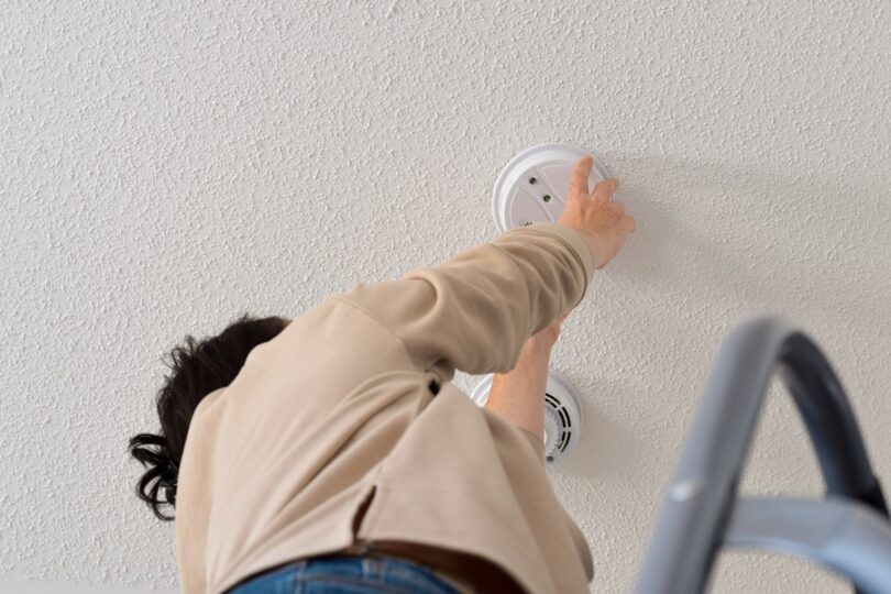 A person on a ladder installs a smoke detector on a textured white ceiling, emphasizing home safety and maintenance.