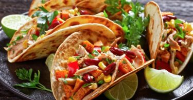 Four tacos filled with leftover chicken, beans, corn, peppers, and fresh cilantro served with lime wedges on a dark plate.