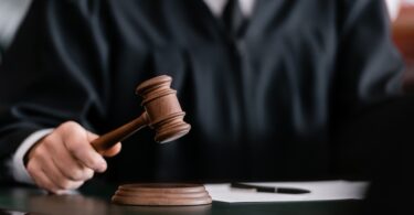 A close-up view shows a judge wearing a black robe holding a wooden gavel. A blank paper with a pen is next to the gavel.
