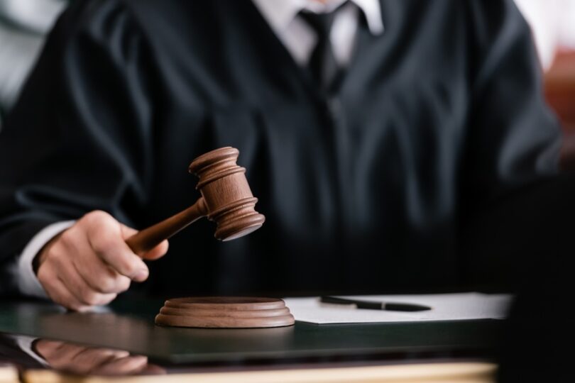 A close-up view shows a judge wearing a black robe holding a wooden gavel. A blank paper with a pen is next to the gavel.