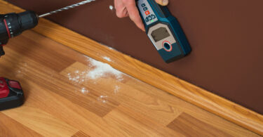 A person holds a stud finder against the brown-painted wall. There is a drill and drill hole dust on the floor nearby.
