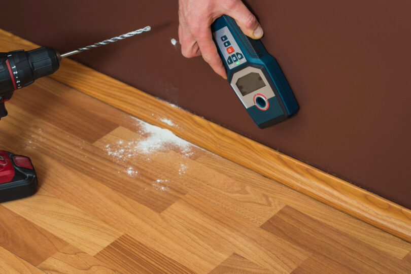 A person holds a stud finder against the brown-painted wall. There is a drill and drill hole dust on the floor nearby.