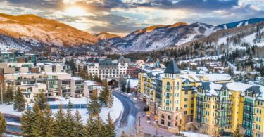 The sun sits over snowy mountains above a city in a valley, including a prominent yellow building and roadways.