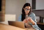 A woman sits at a table in front of a laptop and looks at a smartphone in her hand. The screen is reflected in her glasses.