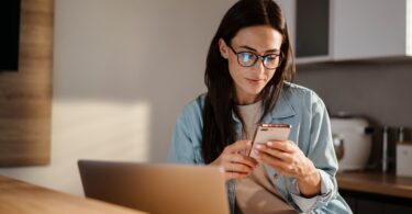 A woman sits at a table in front of a laptop and looks at a smartphone in her hand. The screen is reflected in her glasses.