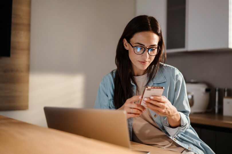 A woman sits at a table in front of a laptop and looks at a smartphone in her hand. The screen is reflected in her glasses.