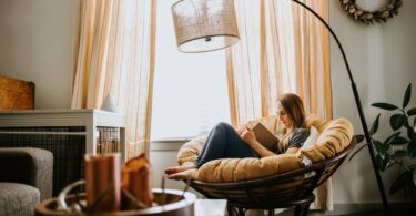 A woman sitting in a large, round chair reading a book. There is a lamp over her and light streaming in through a window.
