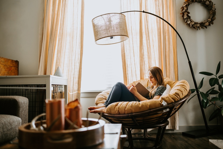 A woman sitting in a large, round chair reading a book. There is a lamp over her and light streaming in through a window.