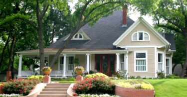 A two-story house with light siding, a dark roof, and a front porch surrounded by a flower garden and lush greenery.
