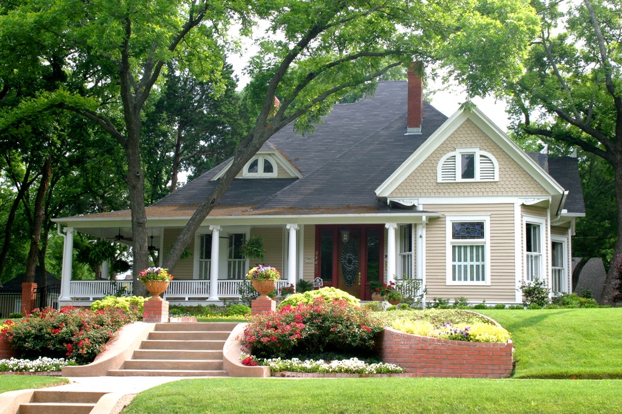 A two-story house with light siding, a dark roof, and a front porch surrounded by a flower garden and lush greenery.