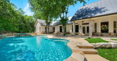 A modern white house with a luxurious pool and hot tub surrounded by mature trees on a clear, sunny day.