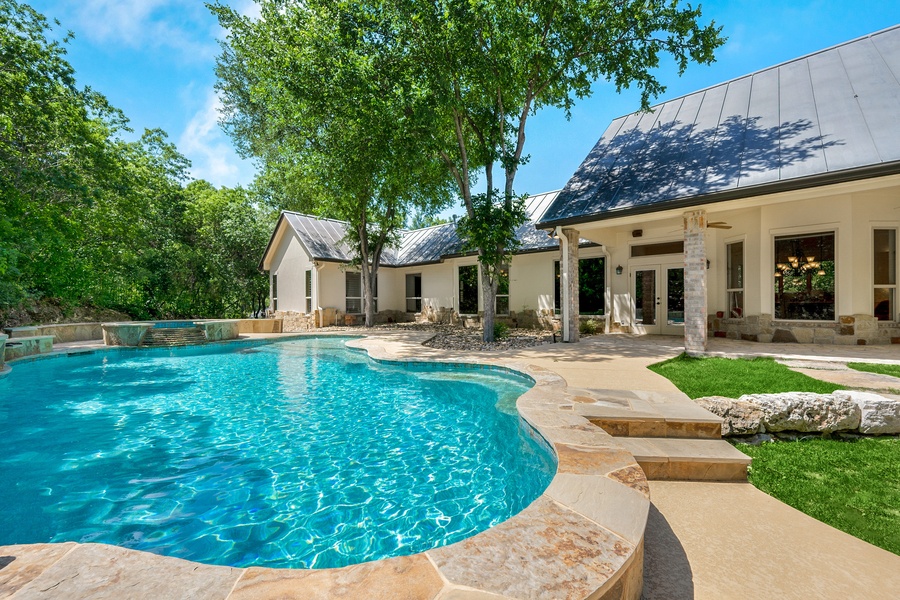 A modern white house with a luxurious pool and hot tub surrounded by mature trees on a clear, sunny day.