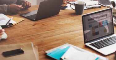 An open laptop that says “Marketing Strategy” on the screen sits on a table with another computer and note sheets.