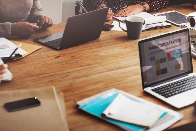 An open laptop that says “Marketing Strategy” on the screen sits on a table with another computer and note sheets.