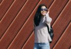A woman in a casual sweater and jeans, wearing sunglasses, poses outdoors against a decorative wooden wall.