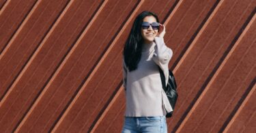 A woman in a casual sweater and jeans, wearing sunglasses, poses outdoors against a decorative wooden wall.