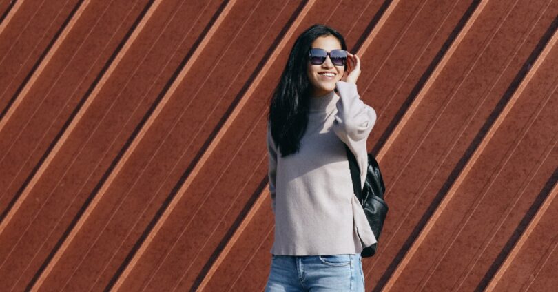 A woman in a casual sweater and jeans, wearing sunglasses, poses outdoors against a decorative wooden wall.