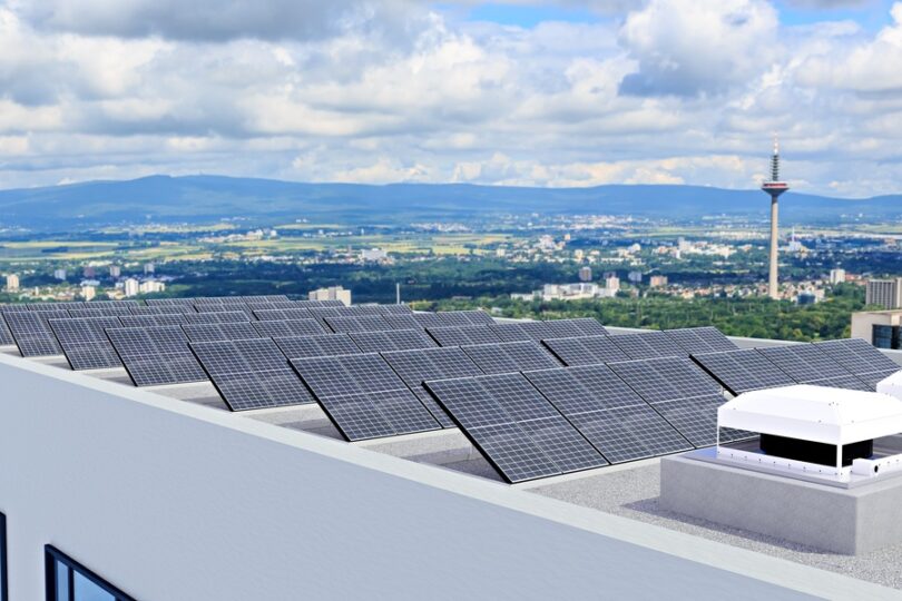 A commercial building with rows of solar panels on the roof, near the HVAC unit. The sky behind the building is cloudy.