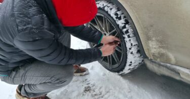 A man wearing multiple layers to stay warm while inflating his car tire. The tire tread is covered in snow.