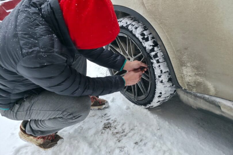 A man wearing multiple layers to stay warm while inflating his car tire. The tire tread is covered in snow.