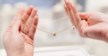 A pair of hands with manicured nails and rings on several fingers holding a necklace with a gold chain and round charm.