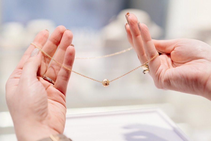 A pair of hands with manicured nails and rings on several fingers holding a necklace with a gold chain and round charm.