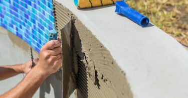 A person spreads ridged mortar on curved concrete with a trowel, near blue mosaic tiles and tools, in an outdoor setting.
