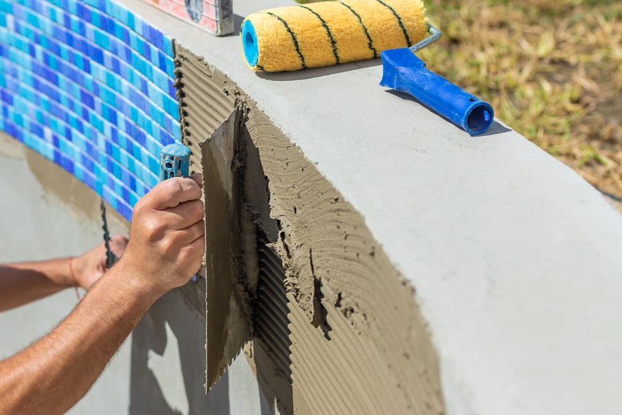 A person spreads ridged mortar on curved concrete with a trowel, near blue mosaic tiles and tools, in an outdoor setting.