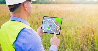 A land inspector looks out over a plot of overgrown land. He has a tablet in his hand depicting housing zones.