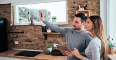 Two people standing in a kitchen at home. One person is holding a pen and clipboard while gesturing in front of them.