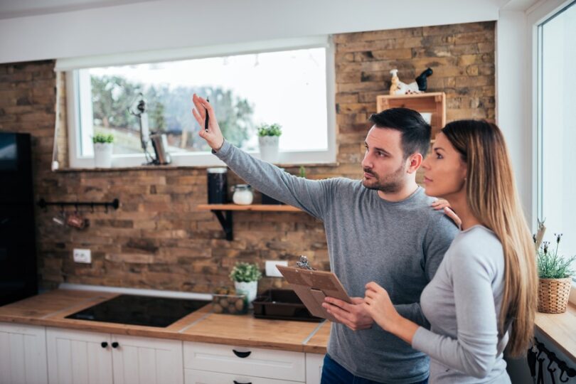 Two people standing in a kitchen at home. One person is holding a pen and clipboard while gesturing in front of them.