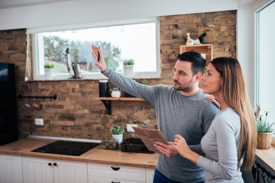 Two people standing in a kitchen at home. One person is holding a pen and clipboard while gesturing in front of them.