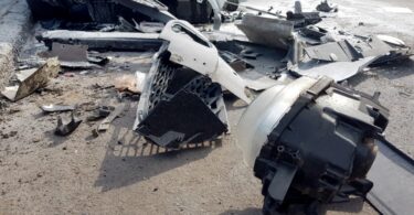 A pile of car parts scattered on the road near a crash site during the daytime. The overturned white vehicle is nearby.