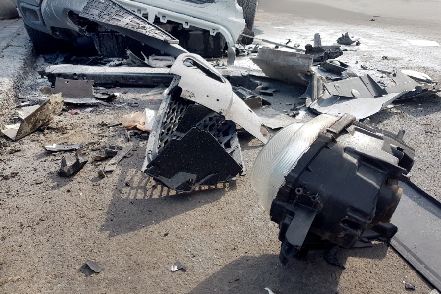 A pile of car parts scattered on the road near a crash site during the daytime. The overturned white vehicle is nearby.