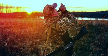 A camouflaged duck hunter blowing a call at sunrise in marsh grass with a shotgun slung over his shoulder.