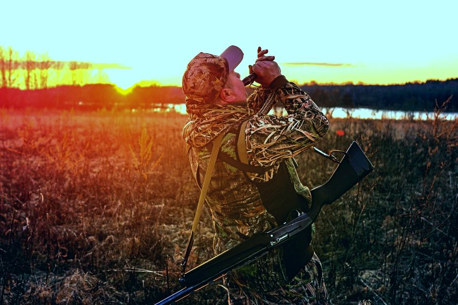 A camouflaged duck hunter blowing a call at sunrise in marsh grass with a shotgun slung over his shoulder.