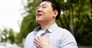 A man stands outside with his head tilted slightly up, his eyes closed, and his mouth parted open. His hand covers his chest.