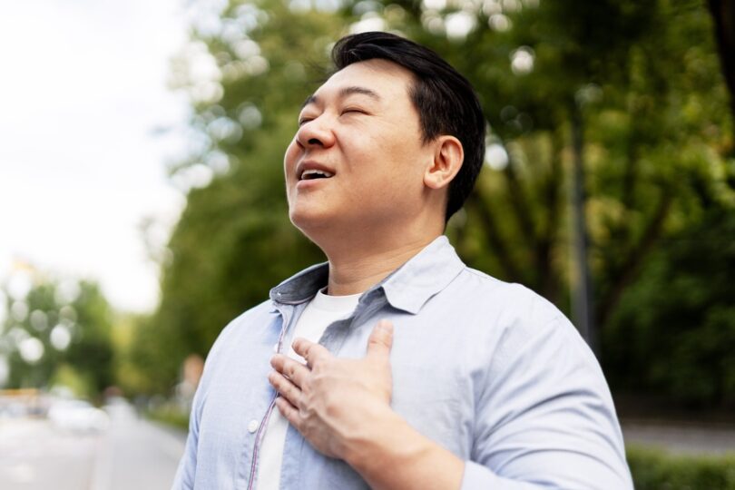 A man stands outside with his head tilted slightly up, his eyes closed, and his mouth parted open. His hand covers his chest.
