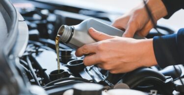 A close-up of an individual pouring golden motor oil from a silver bottle into a car engine during an oil change.
